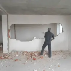 Mężczyzna z młotem burzy ścianę nośną, tworząc otwór. Gruz leży na podłodze.