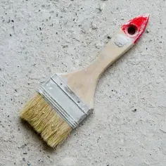 Pędzel z drewnianą rączką i czerwonym zakończeniem leży na szarej, chropowatej powierzchni. Idealny do pytania: czym pomalować beton.