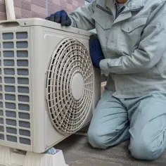 Technik sprawdza klimatyzację w bloku. Urządzenie stoi na zewnątrz, obok ceglanej ściany.