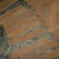 Drewniane deski parkietu, niektóre luźne, czekają na ułożenie. Widać kurz i resztki starej podłogi.