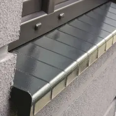 Porysowany parapet z blachy, z widocznymi przetarciami i drobnymi uszkodzeniami, stanowi element zewnętrzny budynku.