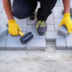 Układanie kostki brukowej z samego piasku i cementu. Ręce w żółtych rękawiczkach układają betonowe elementy.