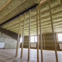 Ocieplenie stropu betonowego wełną mineralną, drewniane elementy konstrukcyjne, okna i grzejniki w pomieszczeniu w trakcie budowy.