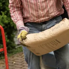 Mężczyzna w rękawicach niesie worek cementu. Zastanawia się, ile piachu na worek cementu 25 kg będzie potrzebne do jego projektu.