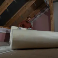 Mężczyzna w rękawiczkach przygotowuje się do ocieplenia dachu od wewnątrz, rozwijając dużą rolkę materiału izolacyjnego.