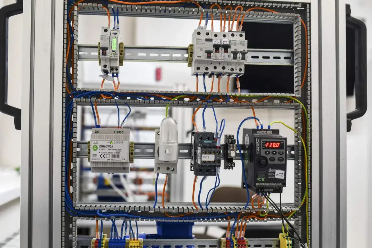 Panel sterowania z elementami do nauki: kurs elektryka od podstaw. Widoczne wyłączniki, przekaźniki, zasilacz i falownik.