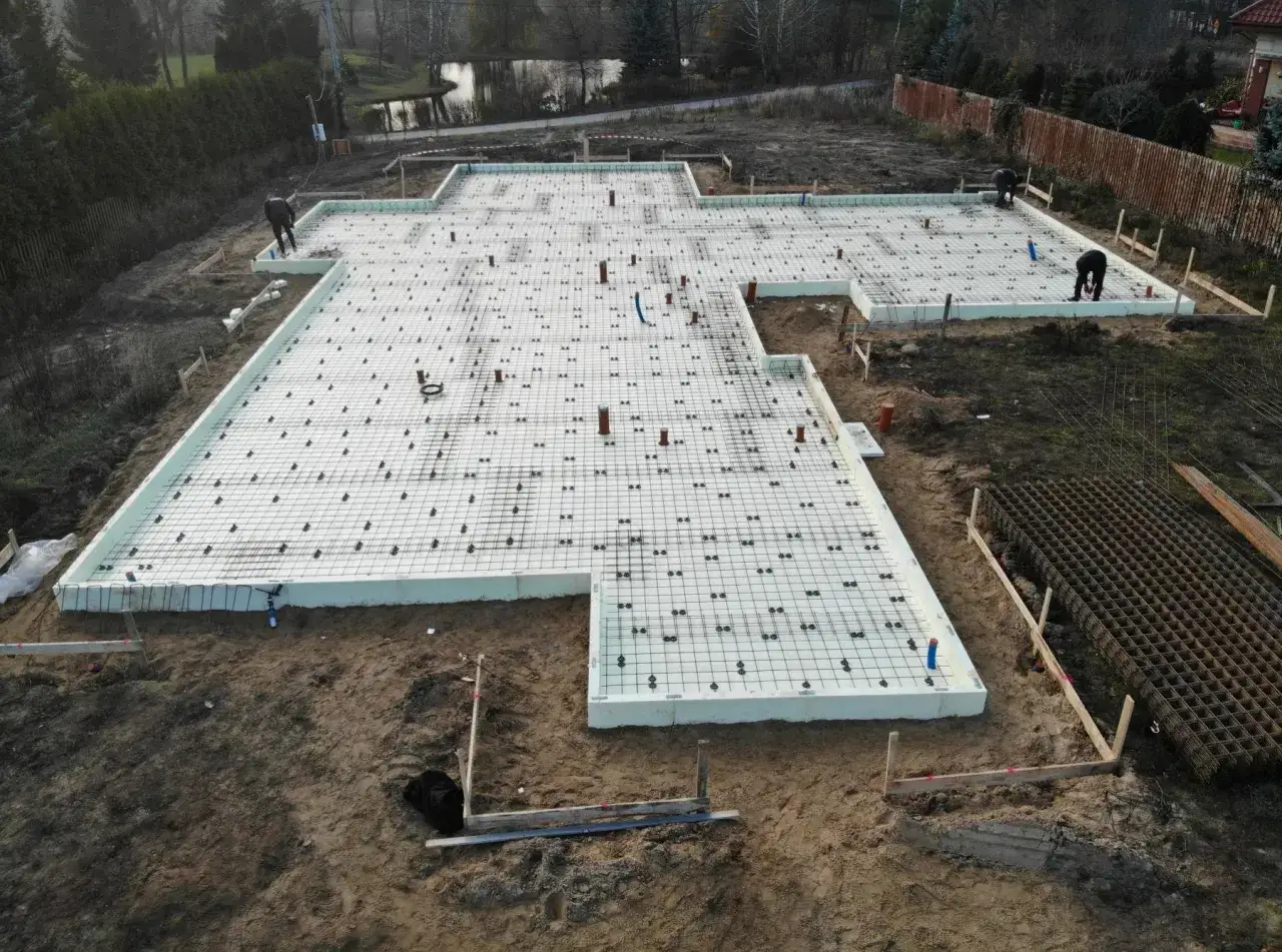 Prace budowlane: przygotowanie pod wylewkę pod garaż. Na placu budowy widać fundamenty z izolacją i zbrojeniem, gotowe do zalania betonem.