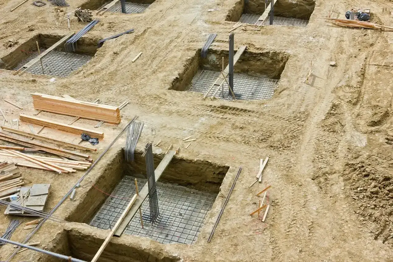 Plac budowy z wykopami pod fundamenty, zbrojeniami i drewnem. Trwa wzmacnianie fundamentów pod przyszły budynek.