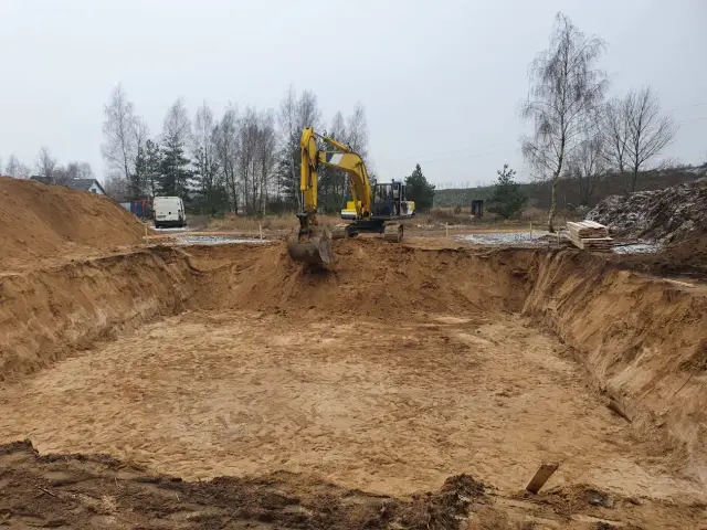 Koparka pracuje przy wykopie, tworząc dół przypominający kurzawkę. W tle widać drzewa i budynki.